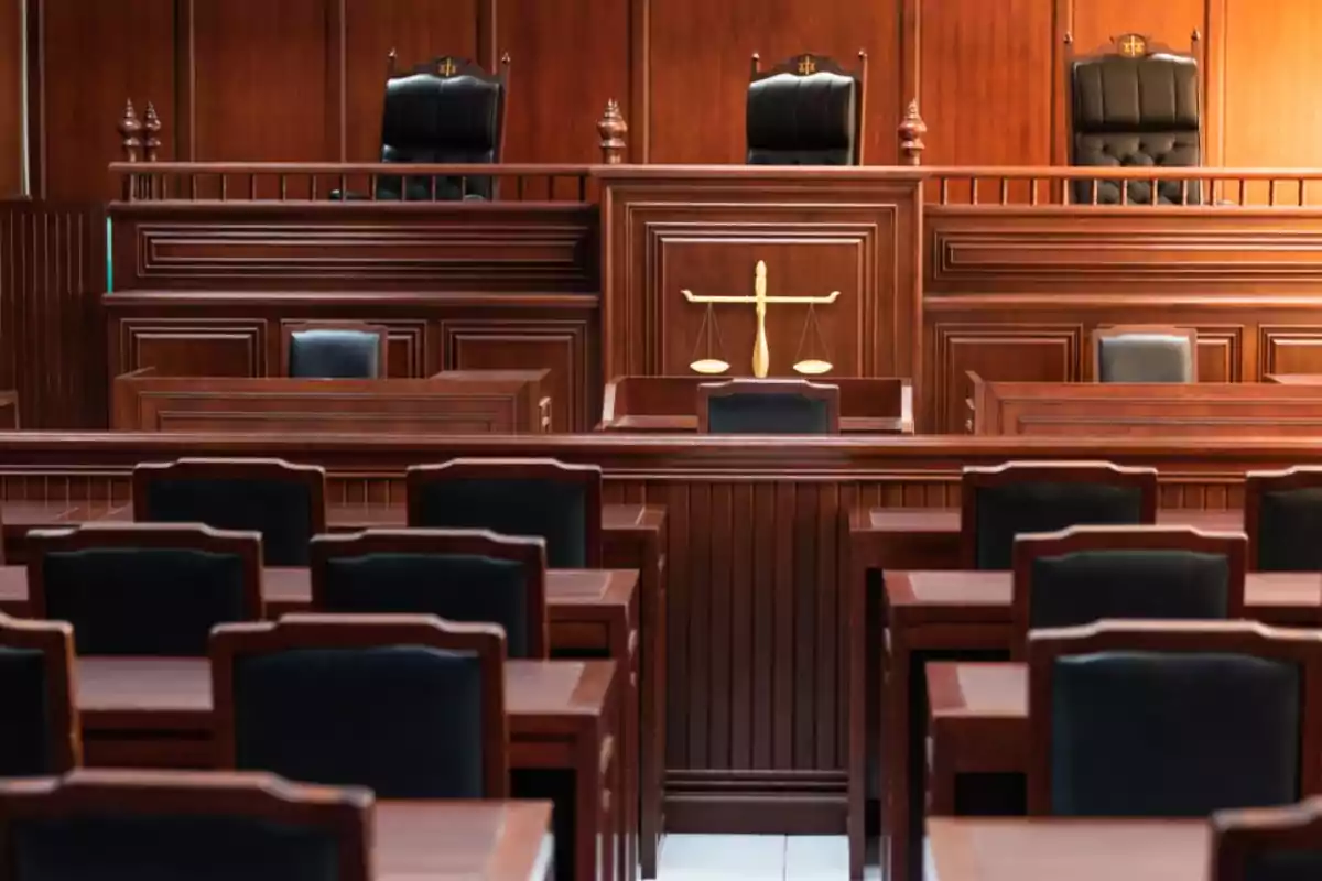 Sala de tribunal vacía con muebles de madera y sillas negras al frente y al fondo una balanza dorada sobre el estrado principal Sala de tribunal vacía con muebles de madera y sillas negras al frente y al fondo una balanza dorada sobre el estrado principal