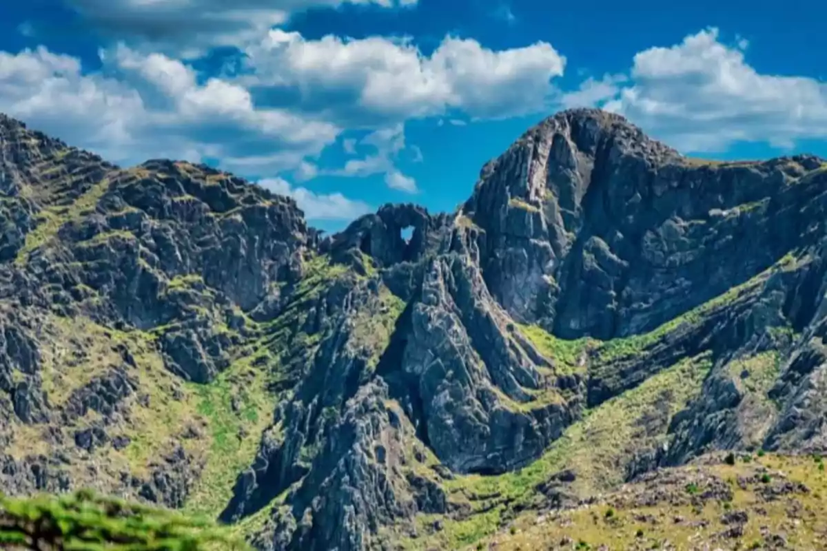 Montañas rocosas con un agujero en forma de corazón bajo un cielo azul con nubes.