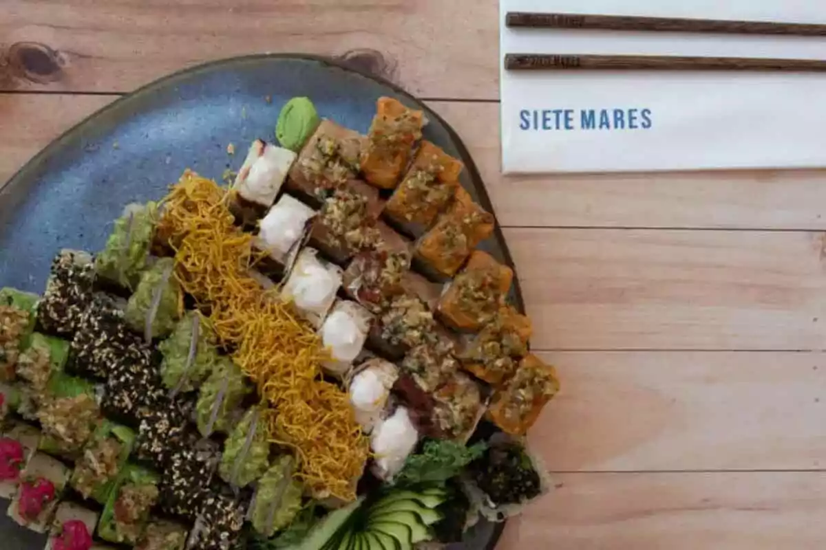 Assorted sushi platter with different types of rolls and side dishes on a wooden table next to a pair of chopsticks and a napkin with the text Siete Mares Assorted sushi platter with different types of rolls and side dishes on a wooden table next to a pair of chopsticks and a napkin with the text Siete Mares