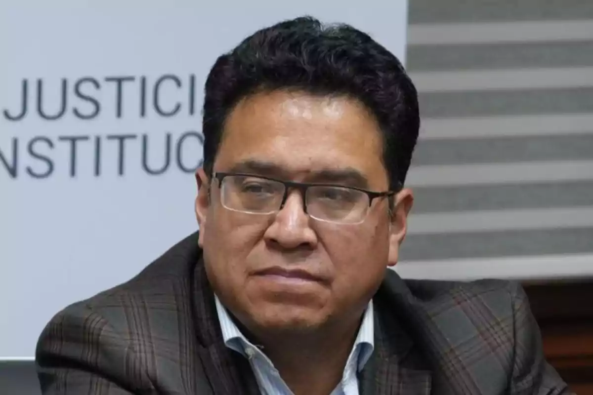 Hombre de cabello corto y gafas con expresión seria usando saco a cuadros y camisa clara sentado frente a un fondo con letras borrosas Hombre de cabello corto y gafas con expresión seria usando saco a cuadros y camisa clara sentado frente a un fondo con letras borrosas