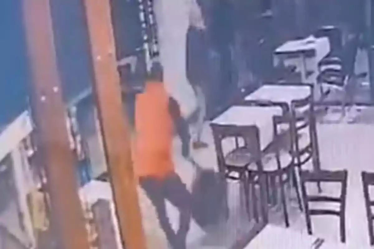 Persona con camiseta naranja agrediendo a otra persona en el interior de un restaurante con varias mesas y sillas vacías
