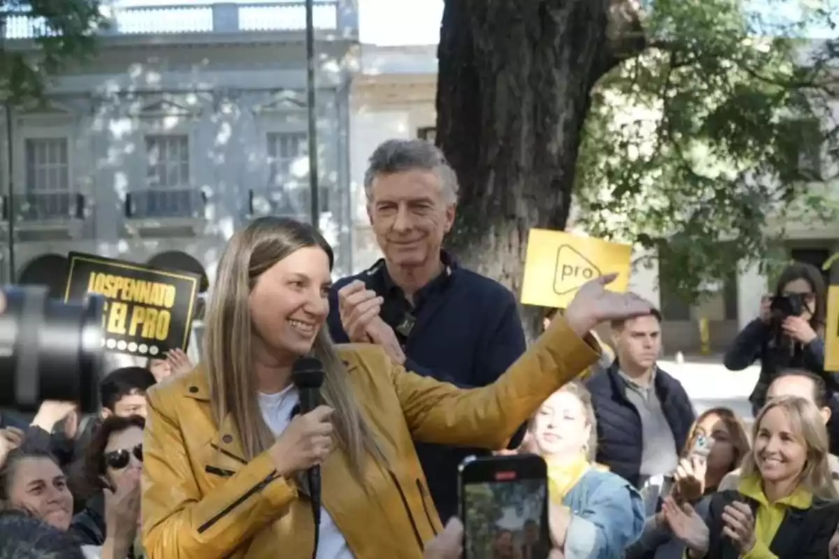 Una mujer sonriente con chaqueta amarilla sostiene un micrófono mientras habla frente a un grupo de personas al aire libre; algunas personas sostienen carteles amarillos. Una mujer sonriente con chaqueta amarilla sostiene un micrófono mientras habla frente a un grupo de personas al aire libre; algunas personas sostienen carteles amarillos.