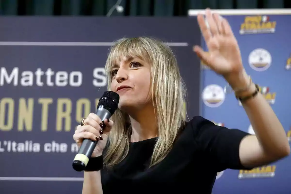 A blonde woman speaking into a microphone during an event with a background that has text and logos.