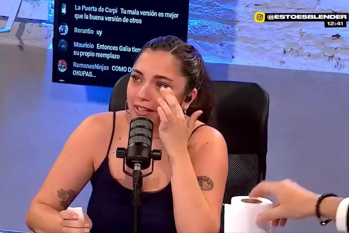 Una mujer en un estudio de grabación está hablando frente a un micrófono mientras se seca las lágrimas con una mano y sostiene un rollo de papel en la otra.