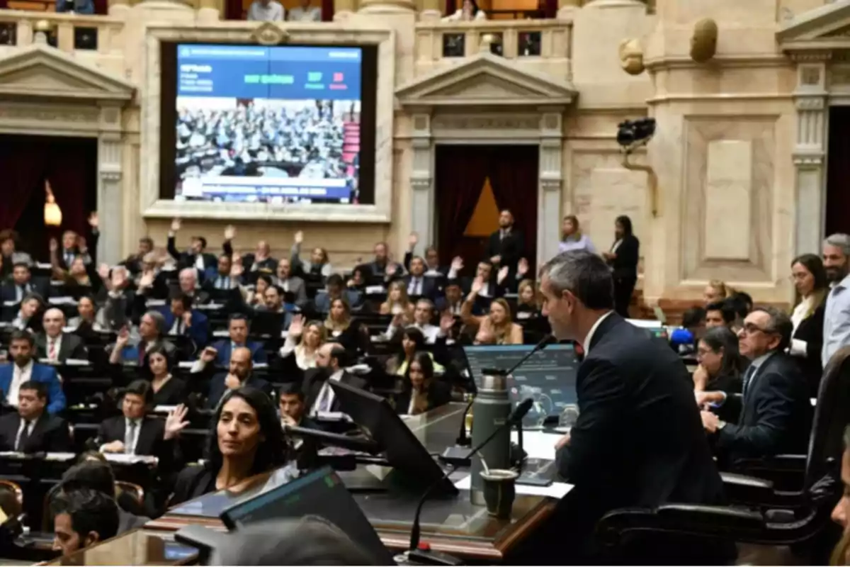 Una sesión parlamentaria con personas levantando la mano en un recinto legislativo, con un gran monitor mostrando información en el fondo.