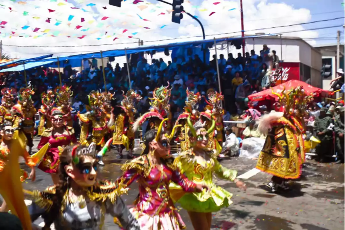 Un grupo de personas disfrazadas con trajes coloridos y máscaras elaboradas participa en un desfile festivo al aire libre mientras el público observa desde las gradas.