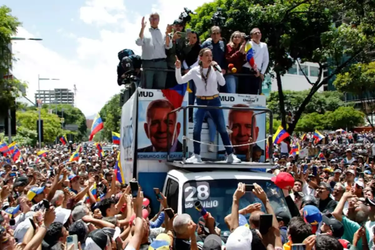 Una multitud de personas en una manifestación política con banderas, mientras una mujer habla desde un camión decorado con carteles de campaña.