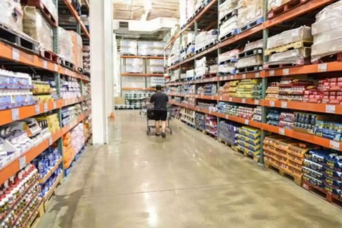 Una persona empuja un carrito de compras por un pasillo de un almacén grande con estantes llenos de productos.