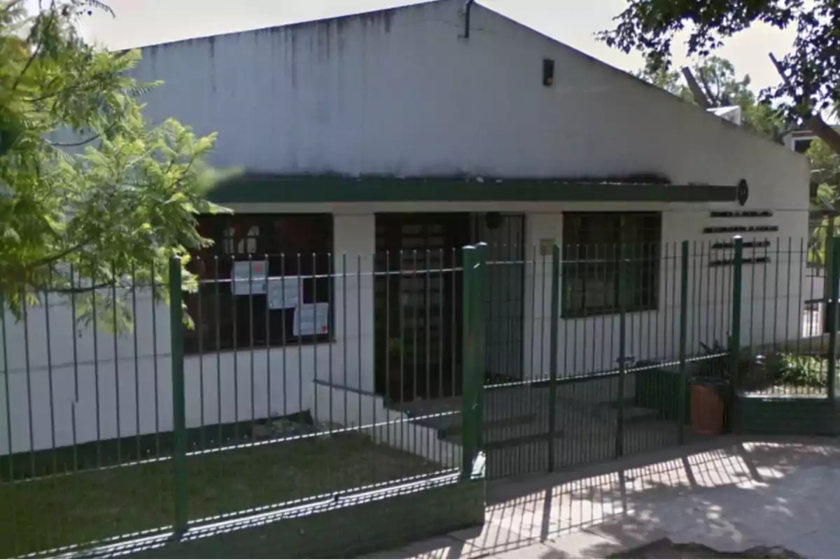 Edificio blanco con techo a dos aguas rodeado de una reja verde y árboles.