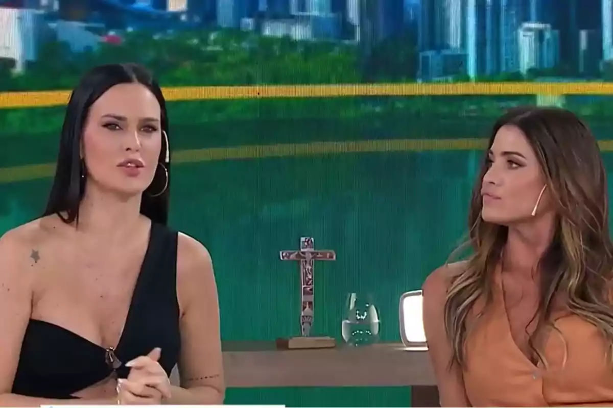 Dos mujeres en un set de televisión con un fondo urbano y una cruz decorativa sobre la mesa.