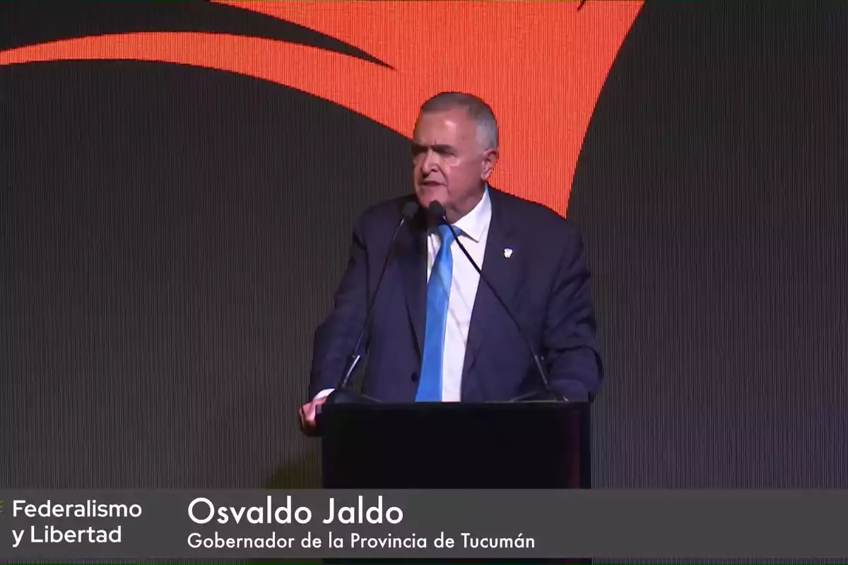 Un hombre de traje y corbata azul habla en un podio con un fondo oscuro y un diseño naranja.