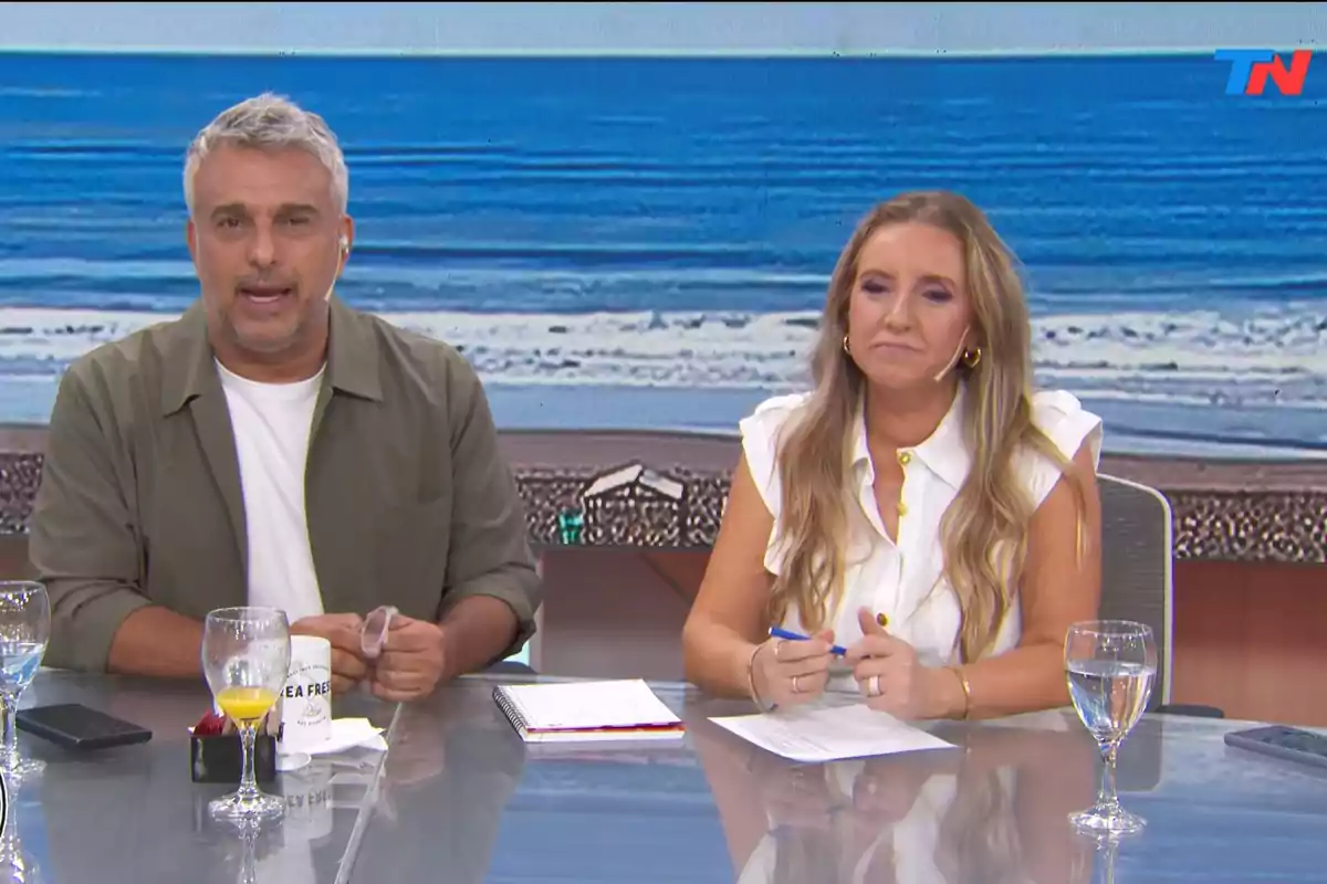 Dos presentadores de televisión están sentados en un estudio con una imagen de playa de fondo, frente a ellos hay una mesa con vasos y papeles.