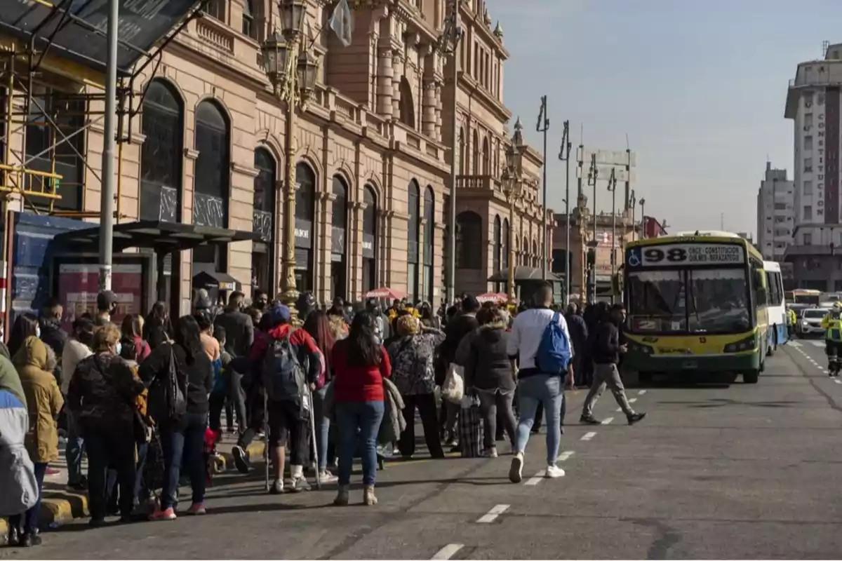 Una multitud de personas espera en una parada de autobús frente a un edificio histórico mientras un autobús se aproxima por la calle.