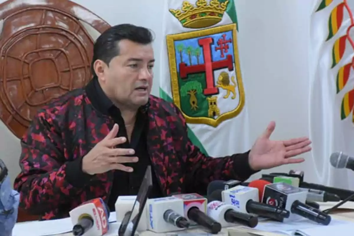 Un hombre hablando en una conferencia de prensa con micrófonos frente a él y un escudo en la pared detrás.