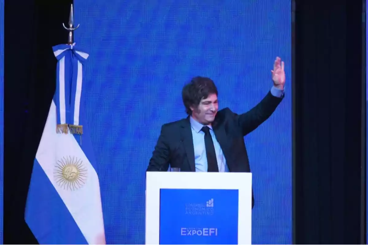 Un hombre en traje oscuro saluda desde un podio con un fondo azul y una bandera argentina a su lado.