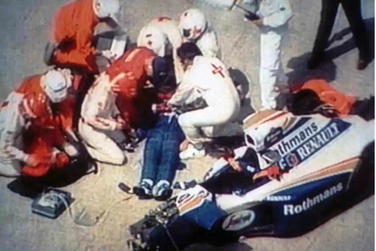 A group of people in emergency uniforms attends to an individual on the ground next to a crashed race car. A group of people in emergency uniforms attends to an individual on the ground next to a crashed race car.