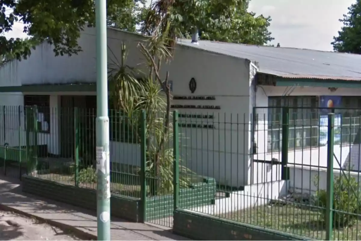 Edificio blanco con techo de chapa rodeado por una reja verde y árboles alrededor.