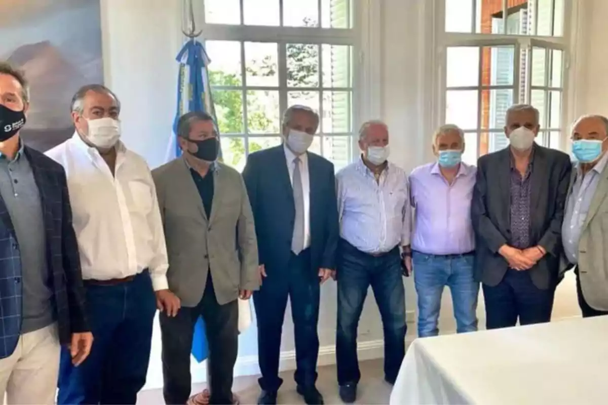 Un grupo de hombres con mascarillas posando juntos en una sala iluminada con ventanas grandes y una bandera argentina al fondo Un grupo de hombres con mascarillas posando juntos en una sala iluminada con ventanas grandes y una bandera argentina al fondo