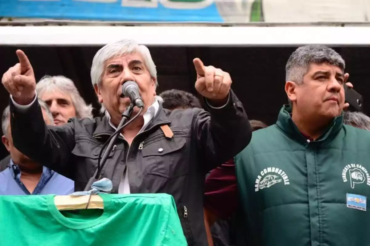 Un hombre hablando por un micrófono en un evento público con otra persona a su lado, ambos rodeados de más personas.