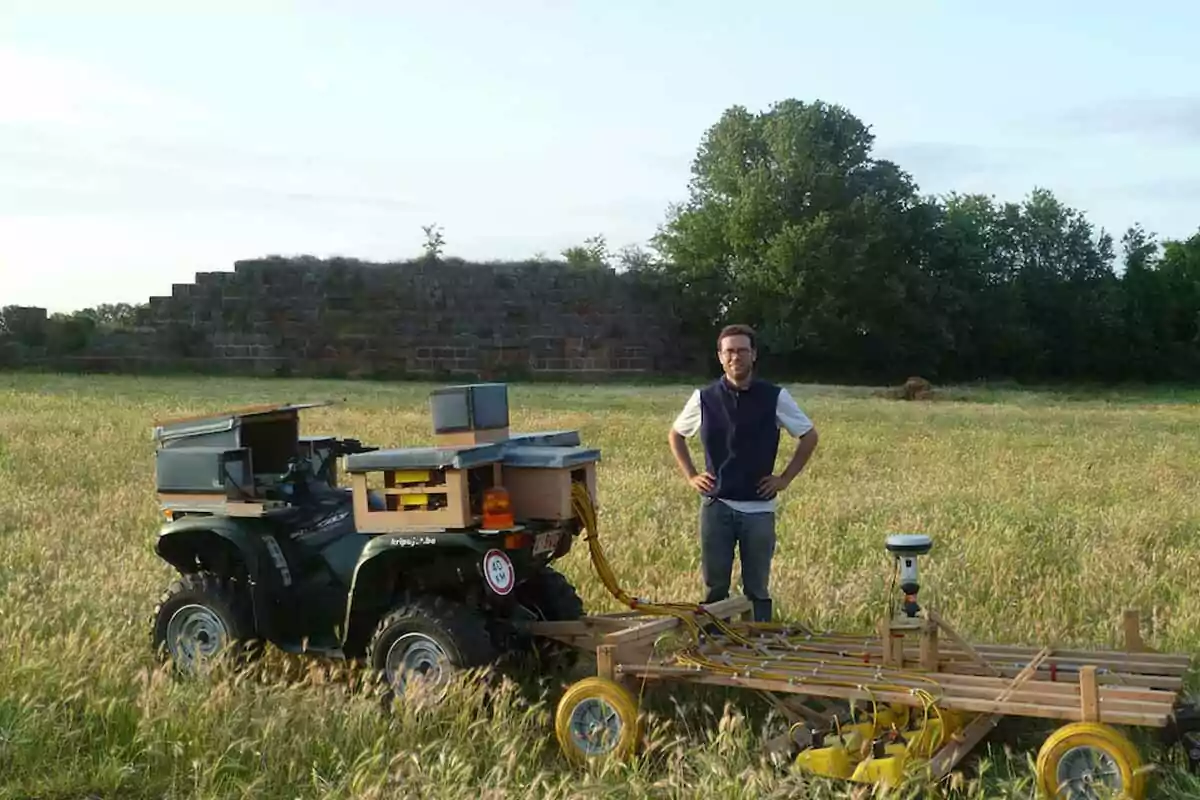 Un hombre de pie en un campo junto a un vehículo todoterreno modificado con equipo especializado y un remolque con ruedas amarillas, con árboles y una estructura de piedra al fondo. Un hombre de pie en un campo junto a un vehículo todoterreno modificado con equipo especializado y un remolque con ruedas amarillas, con árboles y una estructura de piedra al fondo.