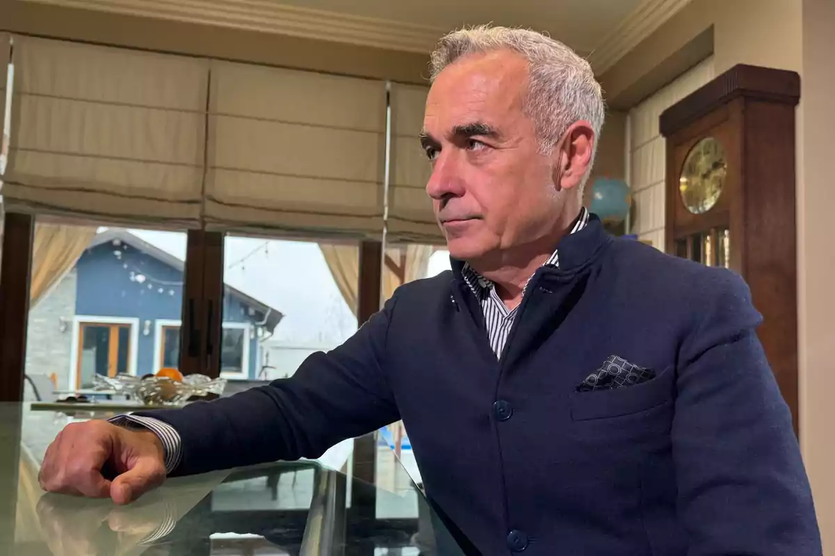 Un hombre de cabello canoso y chaqueta oscura está sentado en una mesa de vidrio en un interior elegante con un reloj de pie y una ventana grande al fondo.