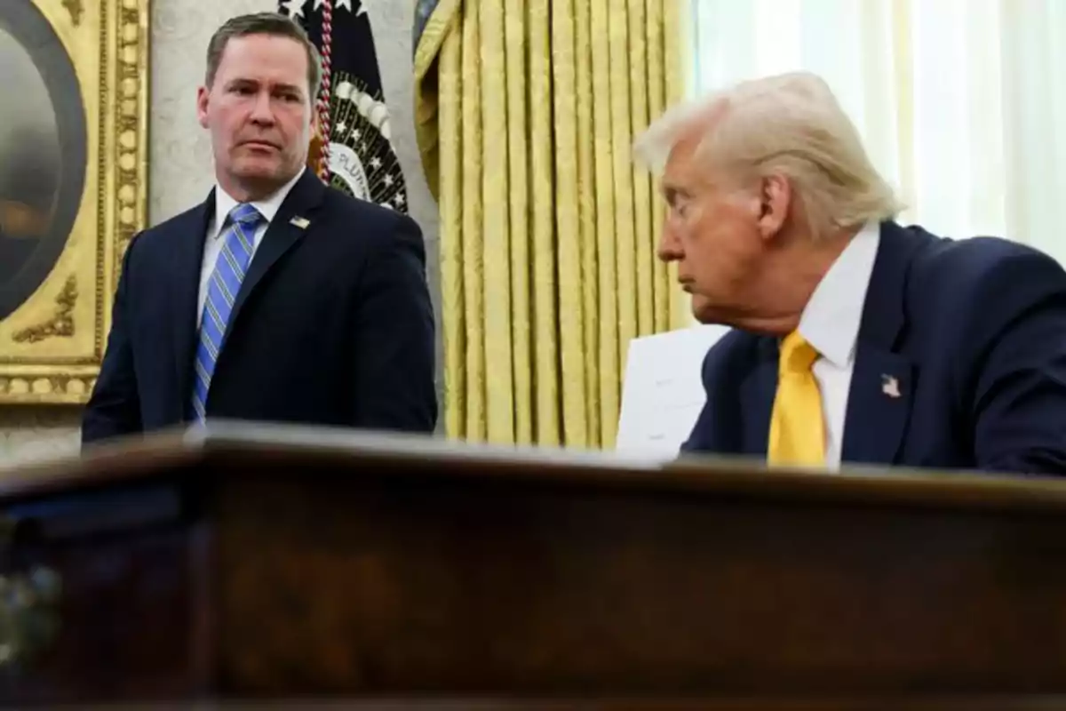 Two men in an office with golden curtains and a desk in the foreground.