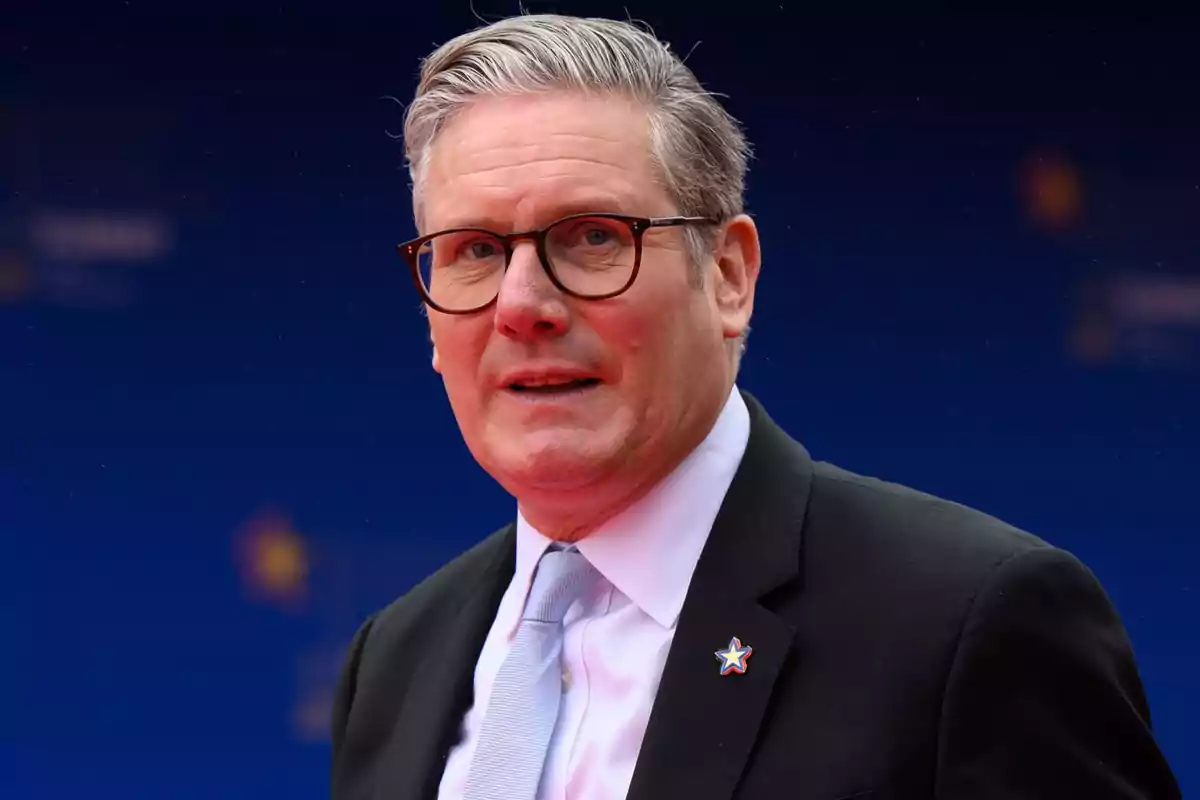 Gray-haired man with glasses wearing a dark suit and light-colored tie against a blue background Gray-haired man with glasses wearing a dark suit and light-colored tie against a blue background