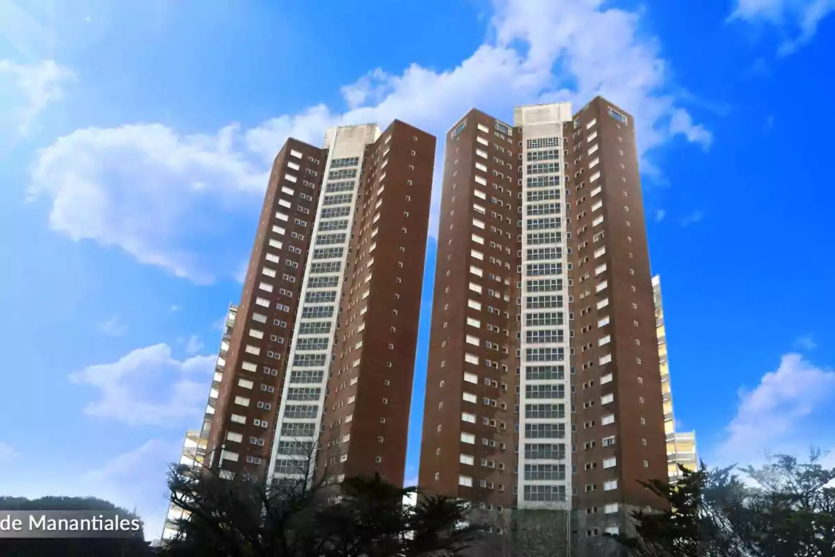 Two tall brick building towers with many windows under a blue sky with some clouds and trees at the bottom Two tall brick building towers with many windows under a blue sky with some clouds and trees at the bottom
