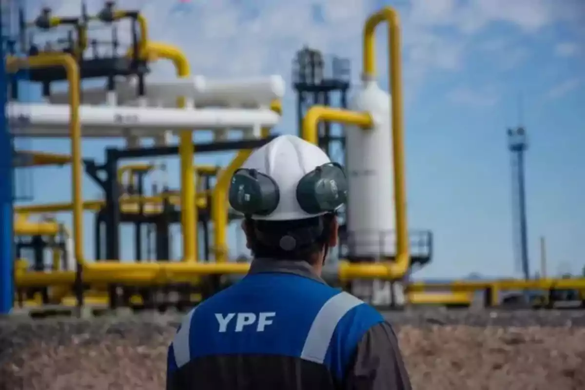 YPF worker with a helmet and hearing protection observes an industrial plant with yellow pipes and metal structures under a clear sky. YPF worker with a helmet and hearing protection observes an industrial plant with yellow pipes and metal structures under a clear sky.