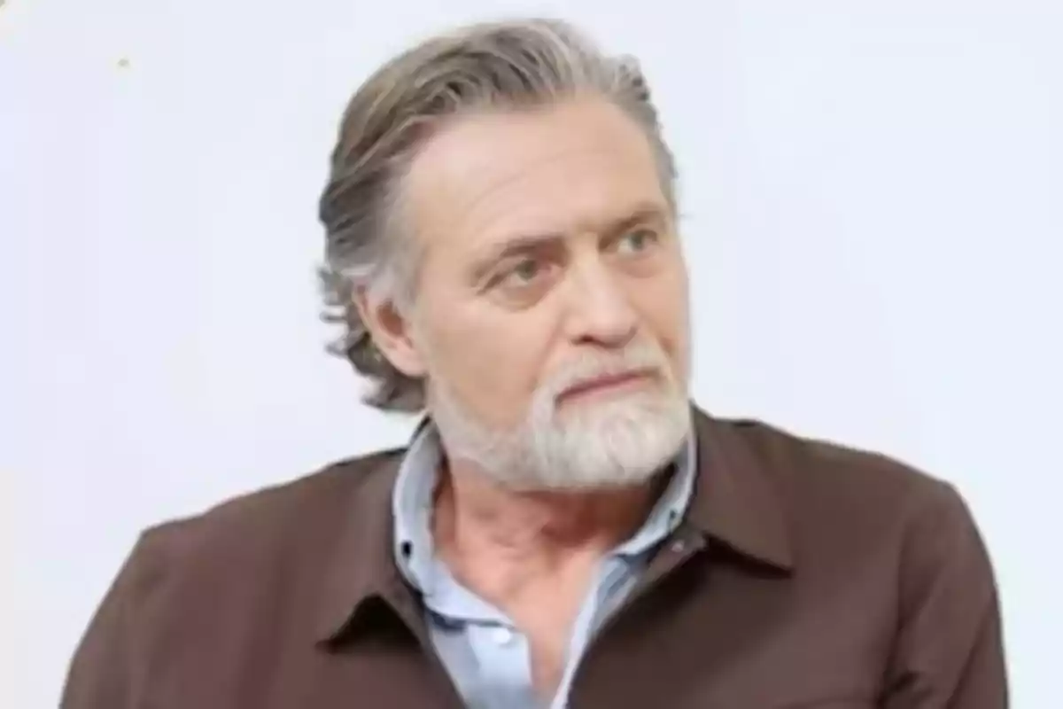 Un hombre de cabello canoso y barba blanca con una chaqueta marrón y camisa azul claro.