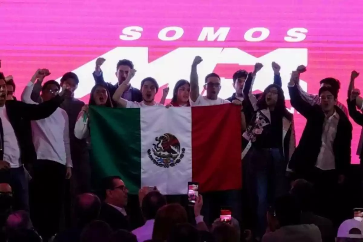 Un grupo de personas sostiene una bandera de México frente a un fondo rosa con letras blancas.