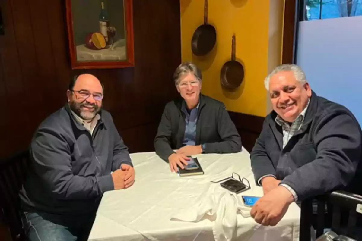 Tres hombres sentados alrededor de una mesa en un restaurante, sonriendo a la cámara, con una pintura y sartenes colgadas en la pared de fondo. Tres hombres sentados alrededor de una mesa en un restaurante, sonriendo a la cámara, con una pintura y sartenes colgadas en la pared de fondo.