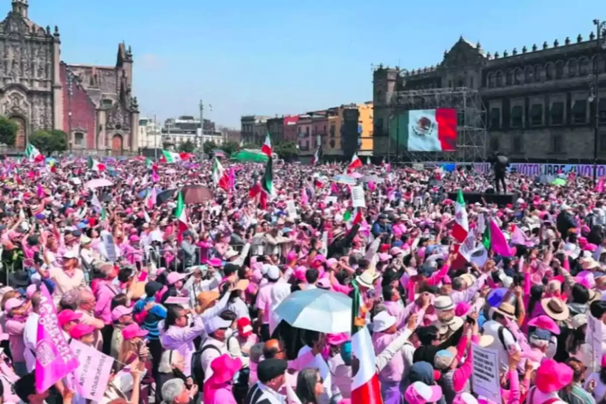 Una multitud vestida de rosa se reúne en una plaza histórica con banderas de México y carteles, mientras un edificio antiguo y una pantalla con la bandera mexicana se ven al fondo. Una multitud vestida de rosa se reúne en una plaza histórica con banderas de México y carteles, mientras un edificio antiguo y una pantalla con la bandera mexicana se ven al fondo.