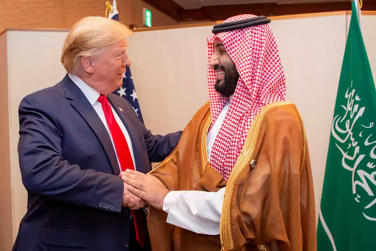Two men shaking hands and smiling, one in a dark suit with a red tie, the other in traditional Arab attire, with flags in the background.