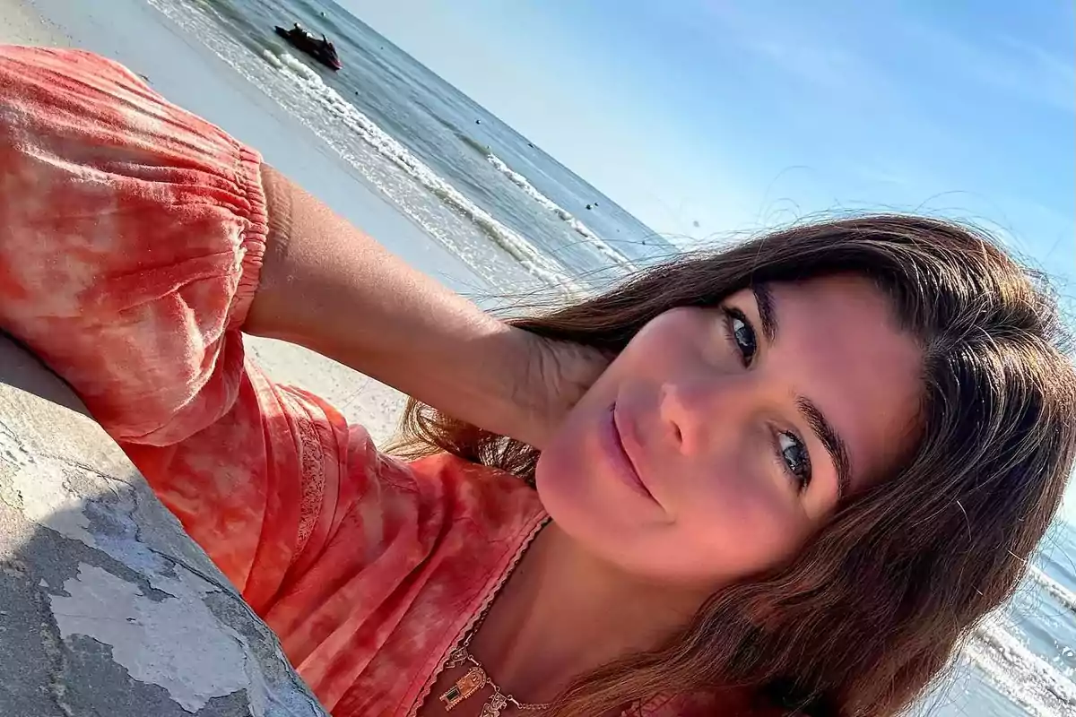 A person in a red blouse smiles while at the beach with the sea in the background.