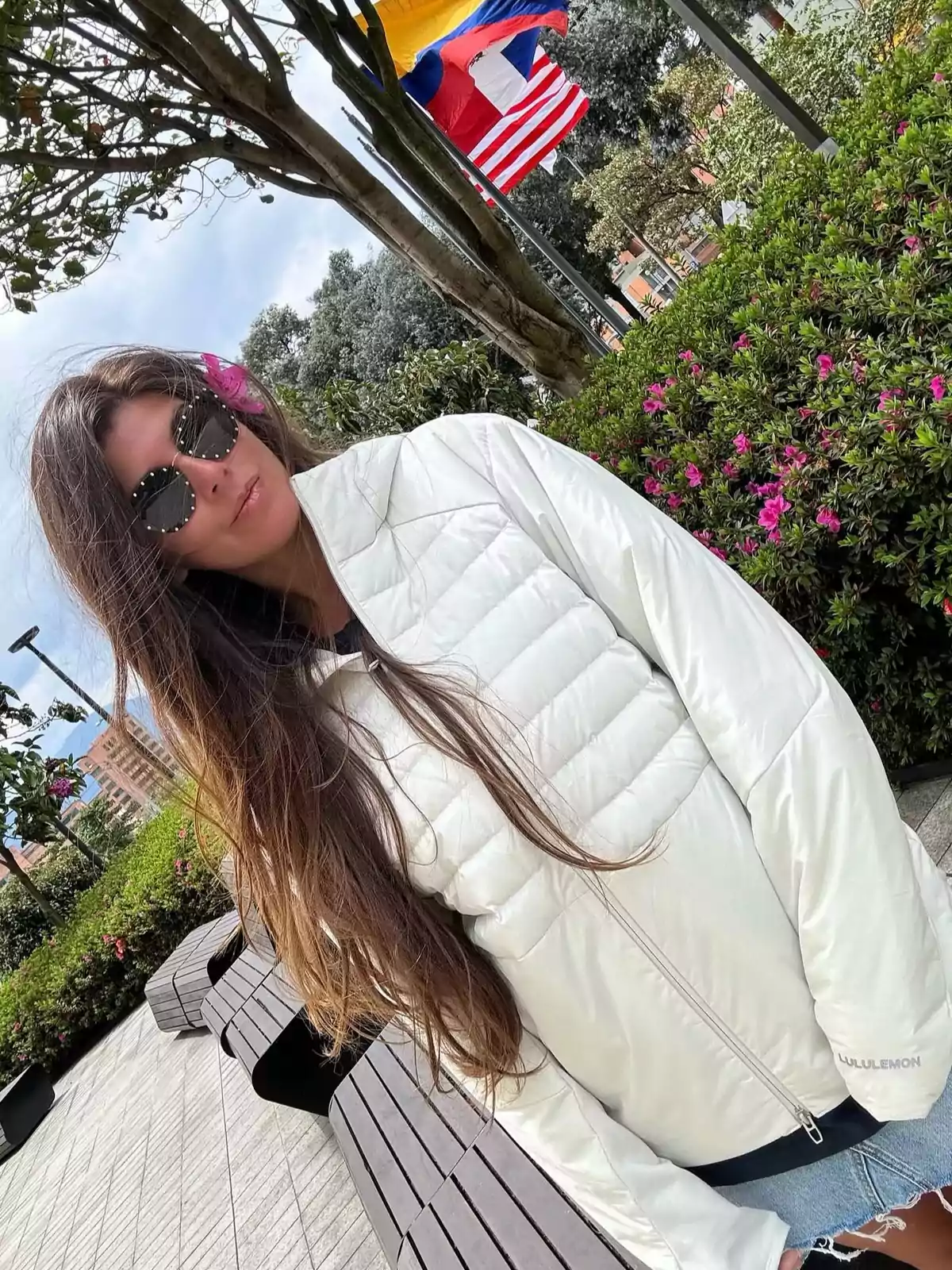 A person with sunglasses and a white jacket is standing in a park with flowers and flags in the background. A person with sunglasses and a white jacket is standing in a park with flowers and flags in the background.