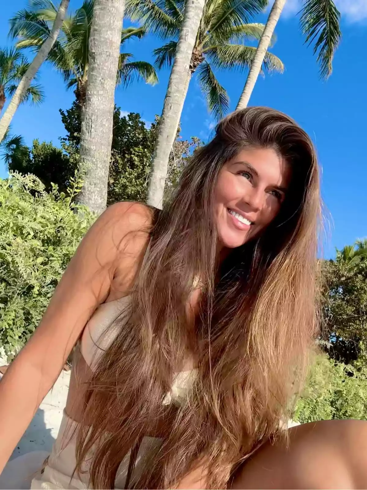 A smiling person with long, loose hair is sitting outdoors surrounded by palm trees and greenery under a blue sky. A smiling person with long, loose hair is sitting outdoors surrounded by palm trees and greenery under a blue sky.
