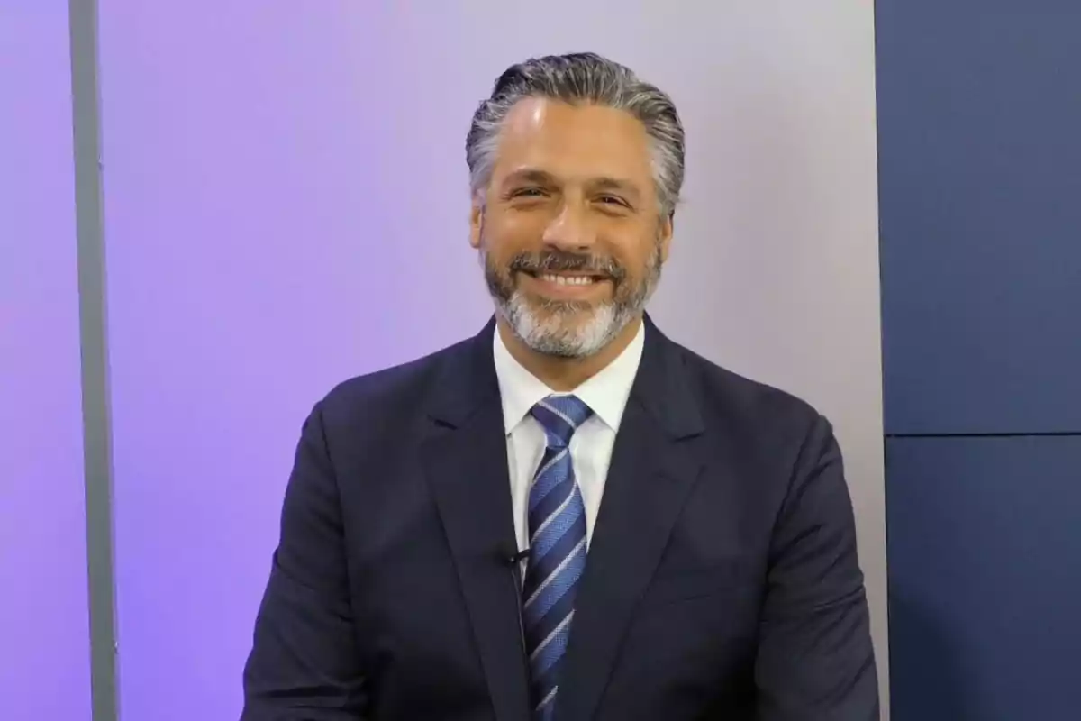 Un hombre con traje oscuro y corbata azul sonriendo frente a un fondo de colores claros. Un hombre con traje oscuro y corbata azul sonriendo frente a un fondo de colores claros.