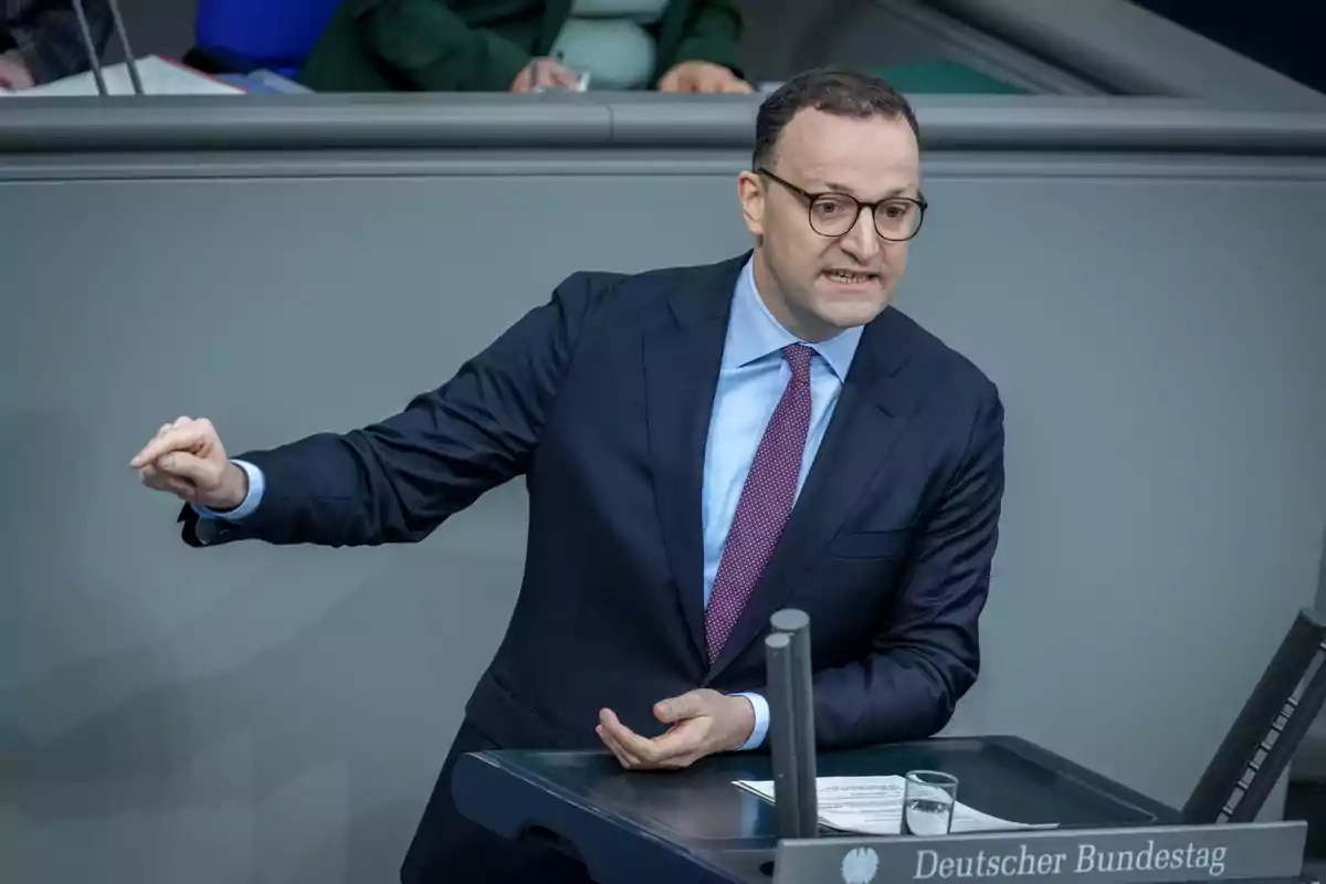 Un hombre con gafas y traje oscuro gesticula mientras habla en un podio en un entorno formal. Un hombre con gafas y traje oscuro gesticula mientras habla en un podio en un entorno formal.