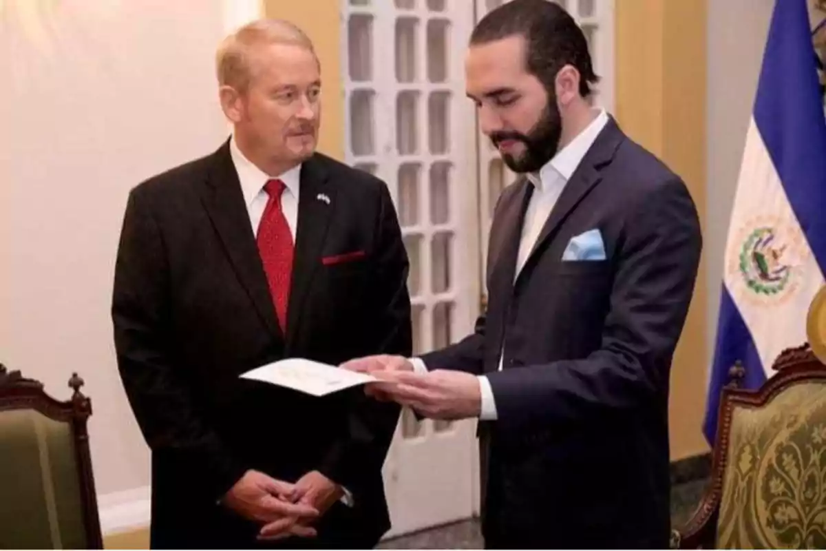 Dos hombres de pie en una sala formal, uno de ellos sostiene un documento mientras el otro observa, con una bandera en el fondo. Dos hombres de pie en una sala formal, uno de ellos sostiene un documento mientras el otro observa, con una bandera en el fondo.