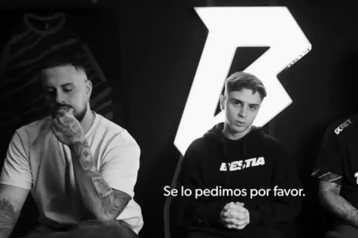 Tres personas sentadas en un entorno oscuro con un logotipo iluminado detrás, una de ellas tiene tatuajes visibles y otra lleva una sudadera con la palabra Tres personas sentadas en un entorno oscuro con un logotipo iluminado detrás, una de ellas tiene tatuajes visibles y otra lleva una sudadera con la palabra