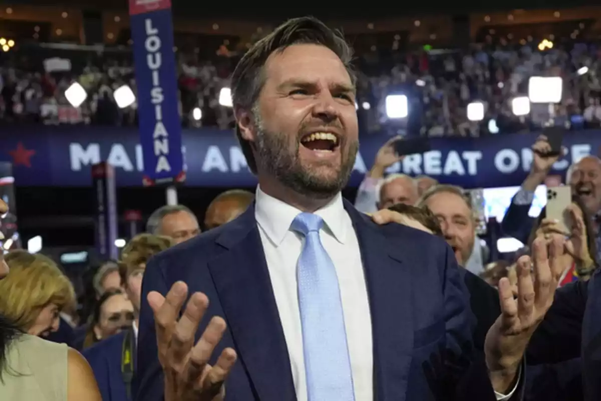 A man in a suit and tie smiles and raises his hands at a crowded event.