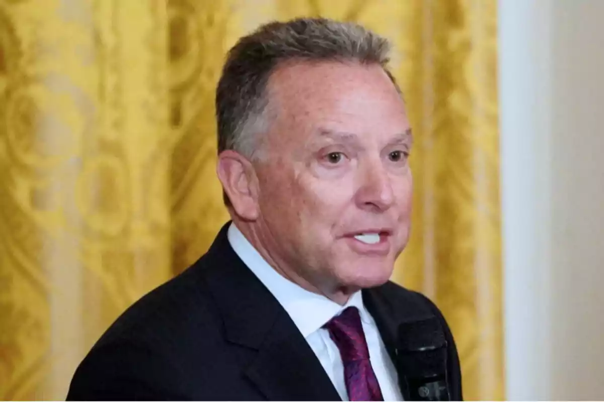 A man in a suit speaking with a microphone in his hand in front of a yellow background.