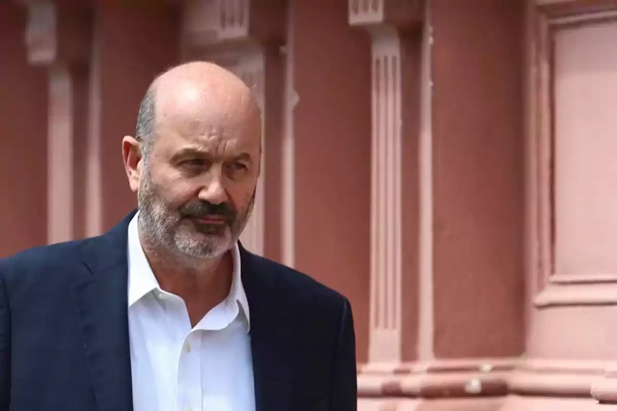 A bearded man in a dark suit is standing in front of a pink building.