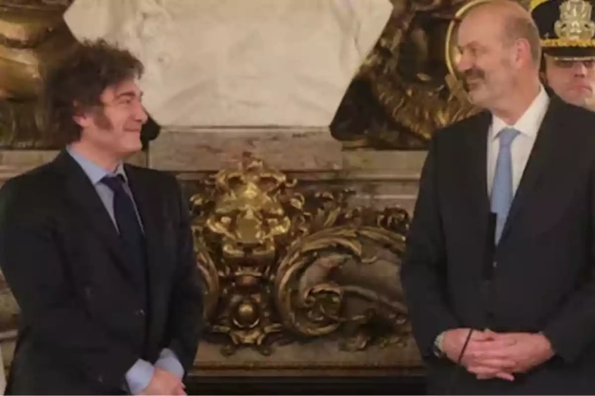 Two men in suits look at each other and smile in an elegant setting with gold decor and a white sculpture in the background.