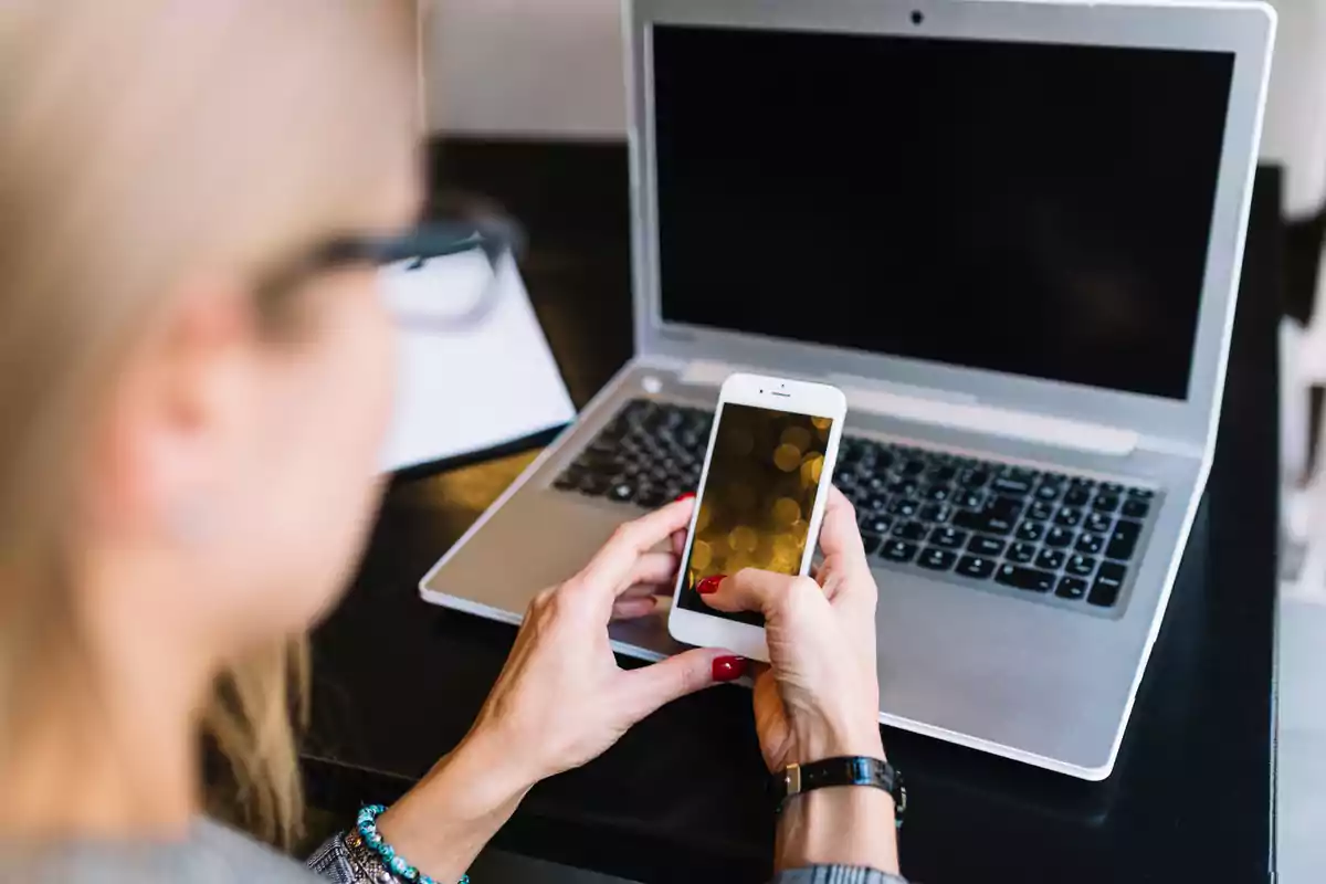 Persona usando un teléfono móvil frente a una computadora portátil en un escritorio.