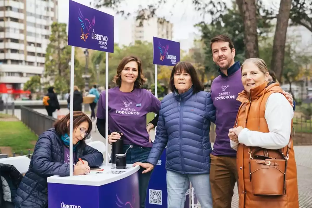 Cinco personas posan en una plaza junto a un stand de campaña política de La Libertad Avanza Córdoba, dos de ellas llevan remeras moradas con el nombre Gabriel Bornoroni y una mujer está escribiendo en una mesa Cinco personas posan en una plaza junto a un stand de campaña política de La Libertad Avanza Córdoba, dos de ellas llevan remeras moradas con el nombre Gabriel Bornoroni y una mujer está escribiendo en una mesa