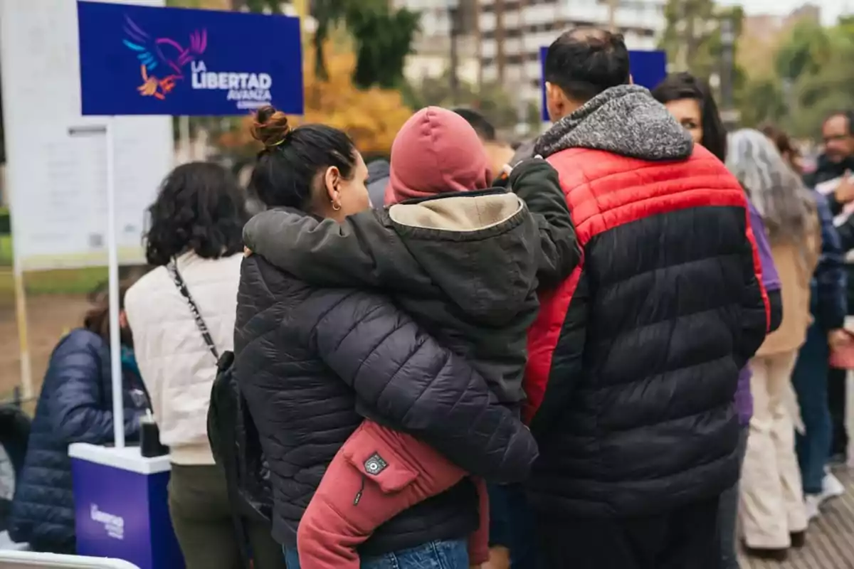 Varias personas con ropa de abrigo hacen fila en un evento al aire libre donde se observa un cartel azul con el texto La Libertad Avanza Varias personas con ropa de abrigo hacen fila en un evento al aire libre donde se observa un cartel azul con el texto La Libertad Avanza