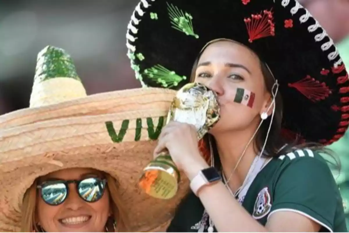Mexican female fans wearing traditional hats celebrate in a stadium; one of them kisses a replica of the World Cup trophy and has a Mexican flag painted on her cheek. Mexican female fans wearing traditional hats celebrate in a stadium; one of them kisses a replica of the World Cup trophy and has a Mexican flag painted on her cheek.