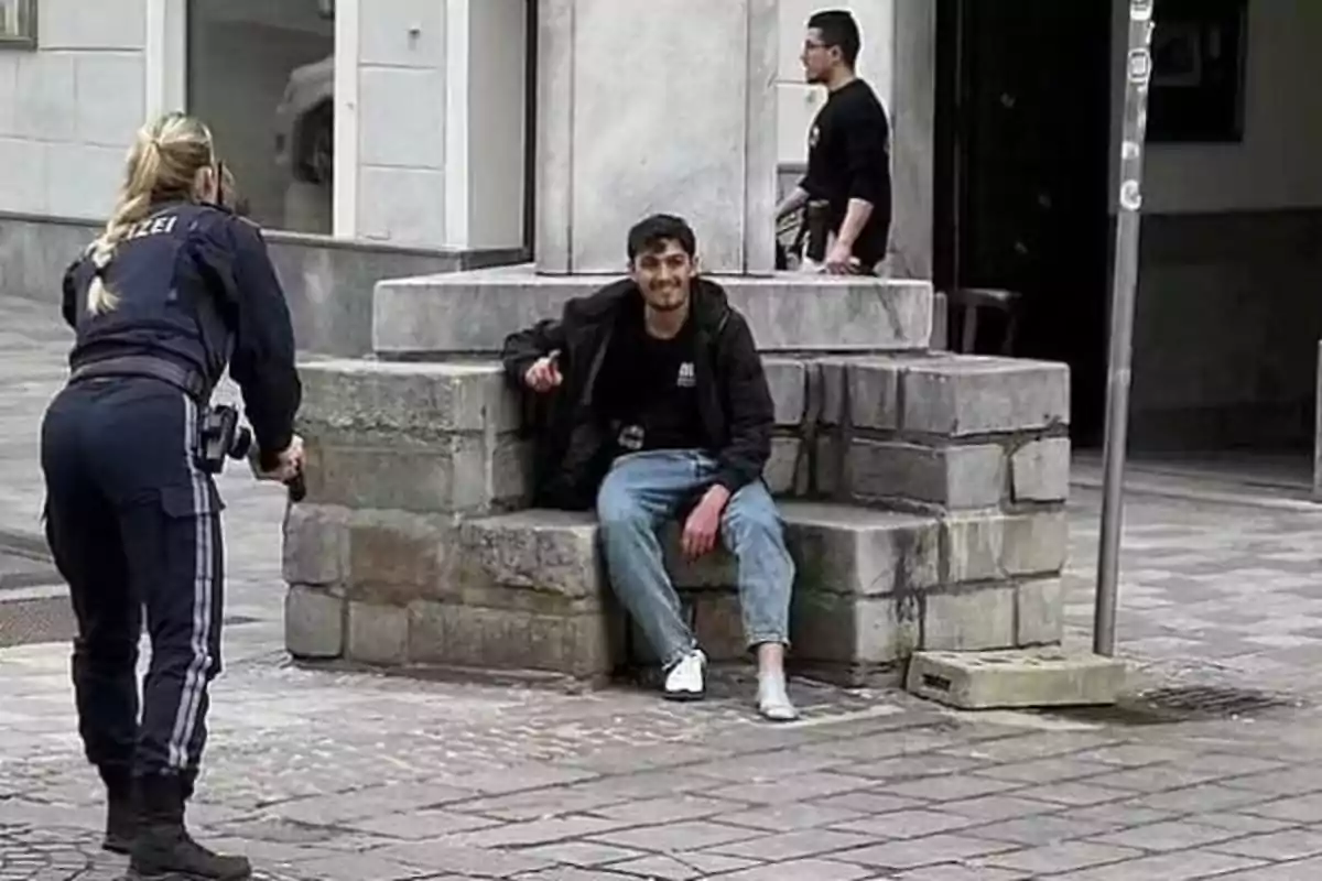 Una oficial de policía apunta con una pistola a un hombre sentado en una estructura de piedra en una calle urbana mientras otra persona camina cerca.